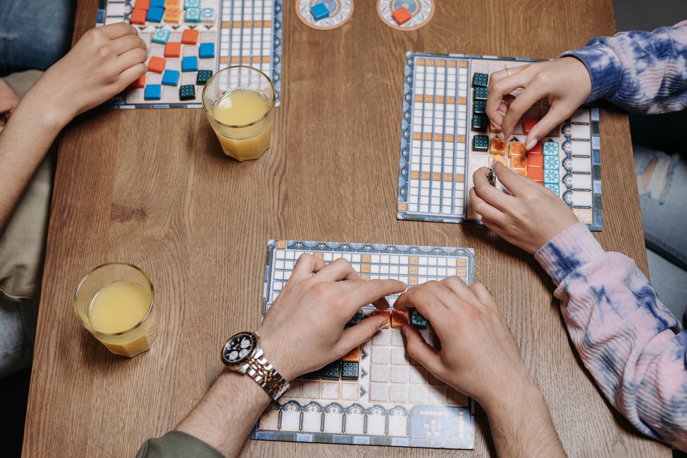 Hands moving tiles on a board game across a wooden table, glasses of juice beside it.
